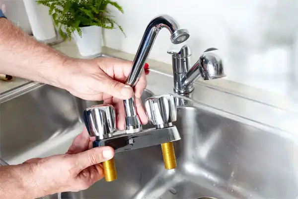Person installing a kitchen faucet with two handles and brass threads, over a stainless steel sink.