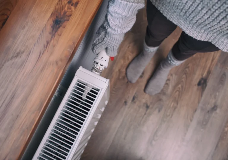 A person wearing knitted gloves and socks adjusts a radiator knob on a wooden floor.