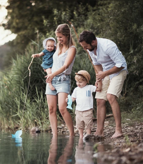 A family of four fishes by a lakeside. The mother holds a baby while the father helps a child with a fishing rod. Trees and grass are in the background.