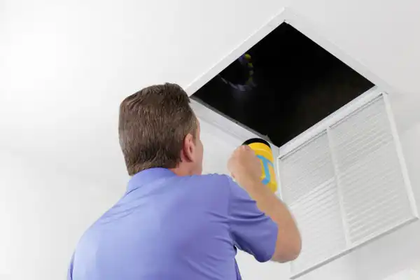 A person in a blue shirt shines a flashlight into an attic access panel on the ceiling.