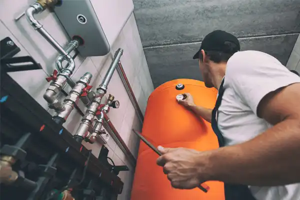 A person in a cap checks an orange water heater tank in a utility room with visible pipes and valves.