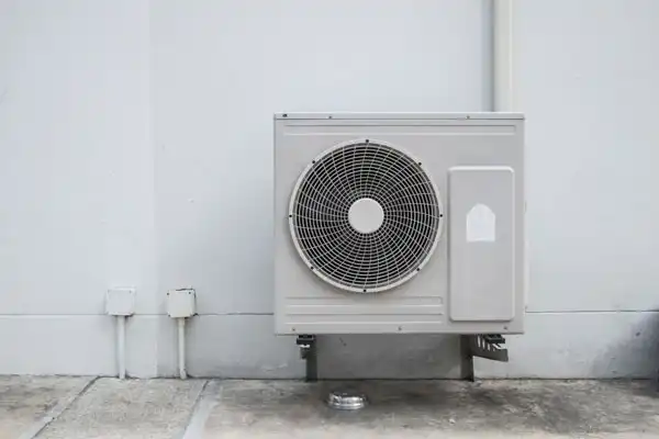 Outdoor air conditioning unit mounted on a white wall with a nearby small pipe and wiring.
