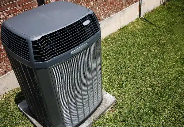 Outdoor central air conditioning unit on a concrete slab, adjacent to a brick wall, surrounded by grass.